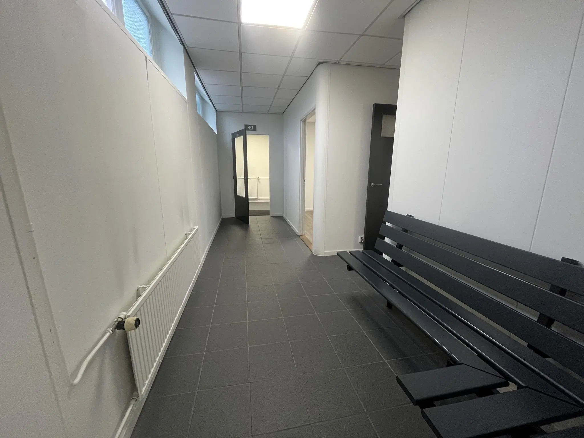 Gang met zwarte tegelvloer, witte muren, een zwarte bank en openstaande deuren in een gebouw aan de Burgemeester Smitsstraat.